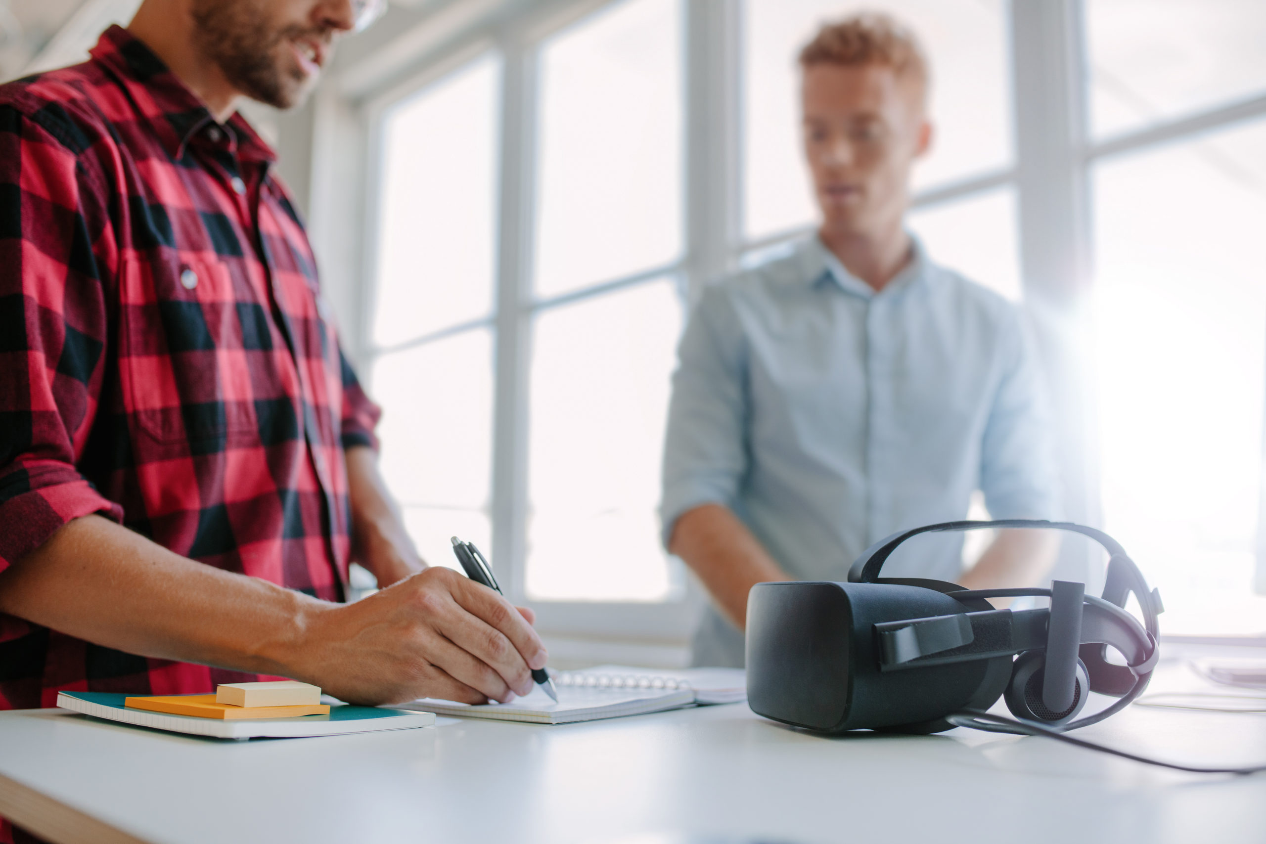 Shot of two young man standing at a table with VR goggles writing on notepad. Developers testing virtual reality glasses in office.